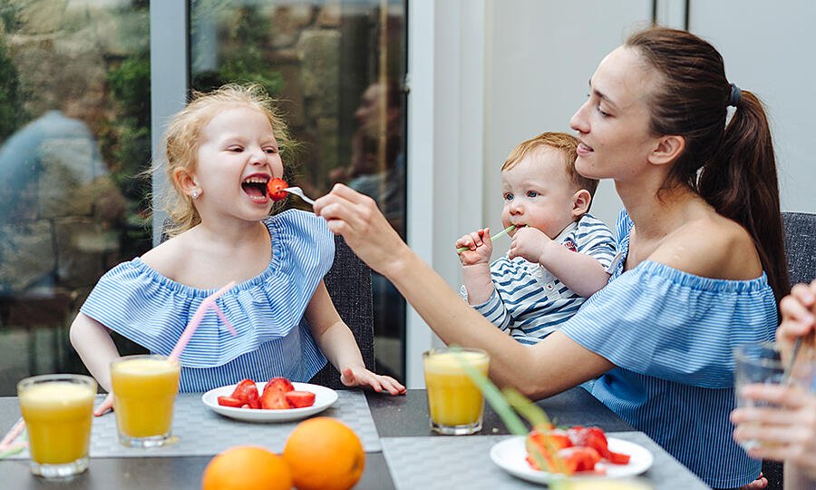 A parent with one child on their hip feeding a second child a plate full of strawberries and presumably putting herself and her meal last because as a parent it be like that sometimes