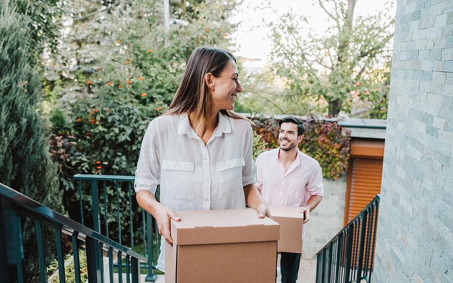 Image of first-time home buyers carrying unmarked boxes up the back stairs of their new home, having a laugh about some indeterminate - probably their matching collars